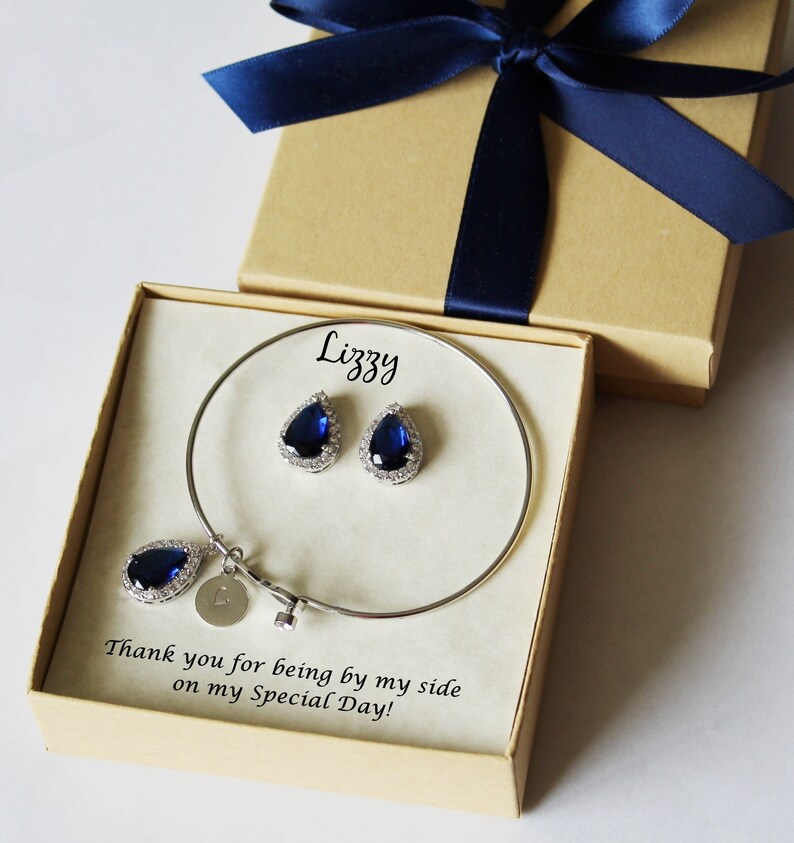 May include: A gift box with a blue ribbon and a personalized message for "Lizzy". Inside the box is a silver bangle bracelet with a teardrop-shaped blue stone charm and a pair of matching earrings.