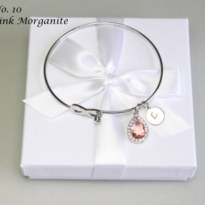 May include: A silver bangle bracelet with a pink morganite gemstone charm and a silver charm with the letter "L" on it. The bracelet is in a white gift box with a white ribbon.