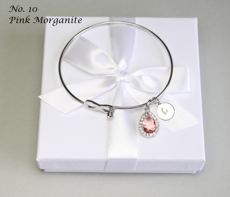 May include: A silver bangle bracelet with a pink morganite gemstone charm and a silver charm with the letter "L" on it. The bracelet is in a white gift box with a white ribbon.