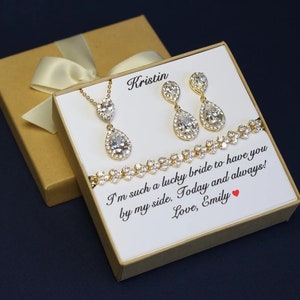 May include: A gift box with a white card inside. The card has a message that reads "I'm such a lucky bride to have you by my side. Today and always! Love, Emily". The box also contains a gold necklace with a teardrop-shaped pendant, a pair of gold earrings with teardrop-shaped pendants, and a gold bracelet with small stones.