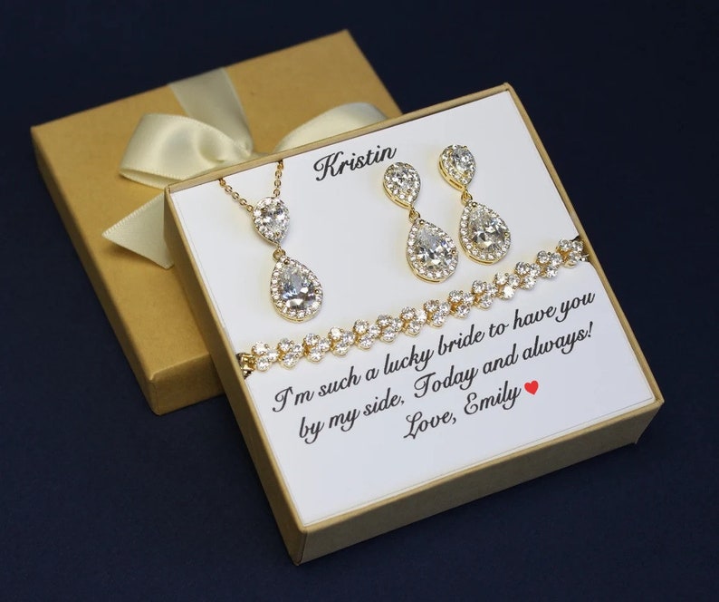 May include: A gift box with a white card inside. The card has a message that reads "I'm such a lucky bride to have you by my side. Today and always! Love, Emily". The box also contains a gold necklace with a teardrop-shaped pendant, a pair of gold earrings with teardrop-shaped pendants, and a gold bracelet with small stones.