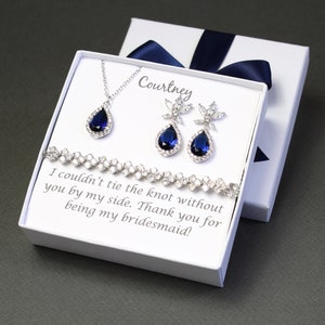 May include: A white gift box with a blue ribbon, containing a blue sapphire necklace, earrings, and bracelet. The box has a personalized message that reads "Courtney I couldn't tie the knot without you by my side. Thank you for being my bridesmaid!"