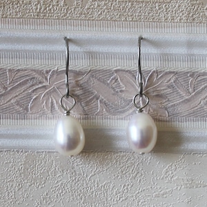 May include: A pair of white pearl drop earrings. Each earring features a lustrous, oval-shaped pearl suspended from a silver-colored hook. The earrings are displayed against a textured, off-white background.