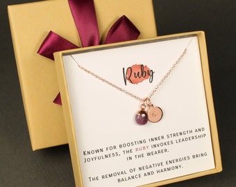 May include: A gold gift box with a red ribbon and a white card inside. The card has a rose gold necklace with a ruby gemstone and a letter charm. The card reads "Ruby. Known for boosting inner strength and joyfulness, the ruby invokes leadership in the wearer. The removal of negative energies bring balance and harmony."