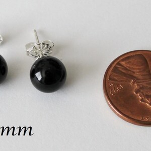 May include: A pair of black onyx stud earrings with silver posts. The earrings are 8mm in diameter and are shown next to a US penny for size comparison.