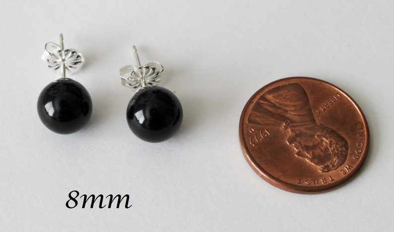 May include: A pair of black onyx stud earrings with silver posts. The earrings are 8mm in diameter and are shown next to a US penny for size comparison.