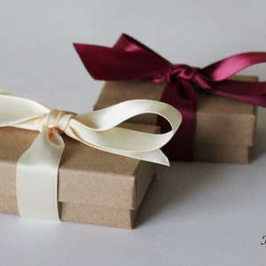 May include: Two small kraft paper boxes with lids, one tied with a cream-colored satin ribbon and the other with a burgundy satin ribbon. The boxes are on a white surface with the text "Kraft box" written below.