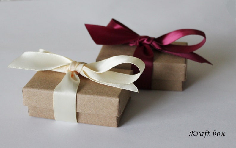 May include: Two small kraft paper boxes with lids, one tied with a cream-colored satin ribbon and the other with a burgundy satin ribbon. The boxes are on a white surface with the text "Kraft box" written below.