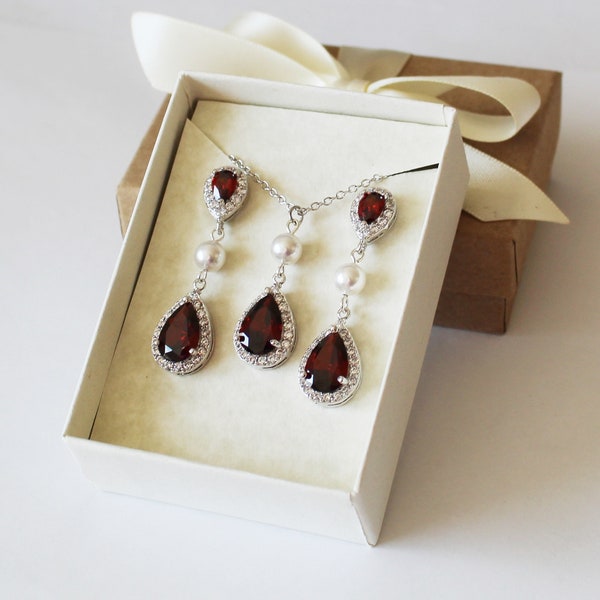 May include: A set of red gemstone earrings and a necklace with pearl accents. The jewellery is displayed in a white box with a brown lid.