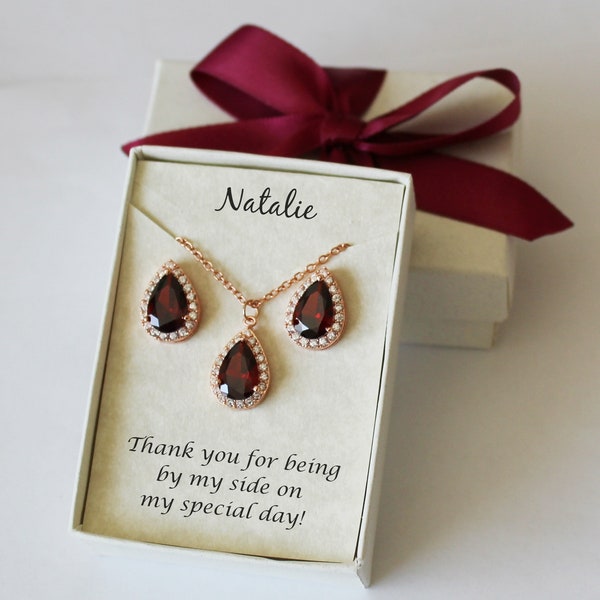 May include: A rose gold necklace and earring set with teardrop-shaped, dark red stones and clear crystal accents. Presented in a gift box with a burgundy ribbon, the card is inscribed with "Natalie" and the message "Thank you for being by my side on my special day!"