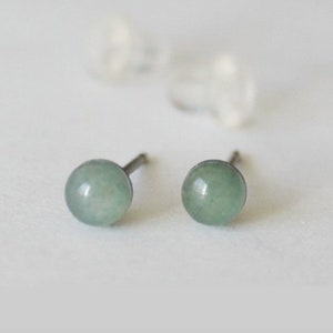 May include: A pair of stud earrings featuring round, pale green gemstones. The earrings have a simple, minimalist design with a post backing. The image also shows two clear earring backs. The earrings are set against a white background.