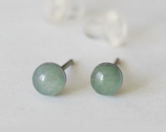 May include: A pair of stud earrings featuring round, pale green gemstones. The earrings have a simple, minimalist design with a post backing. The image also shows two clear earring backs. The earrings are set against a white background.