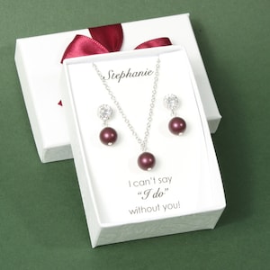Op de afbeelding: Een sieradenset in een witte geschenkdoos met een bordeauxrood lint. De set bevat een ketting en oorbellen met zilveren kettingen en donkerrode parelachtige kralen. De doos heeft de naam "Stephanie" en de tekst "I can't say 'I do' without you!"