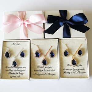 May include: Three gift boxes with blue and pink ribbons. Each box contains a necklace and earrings set with a teardrop-shaped blue stone. The boxes have personalized messages: "Ashley, I couldn't imagine our wedding without you! Thanks for being by my side.", "Thank you for standing by my side. Today and Always!" and "Thank you for standing by my side. Today and Always!"