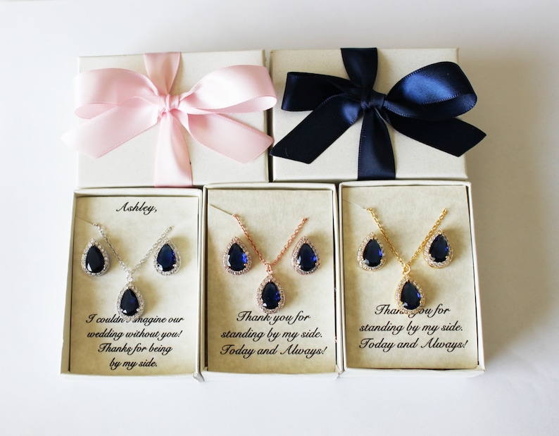 May include: Three gift boxes with blue and pink ribbons. Each box contains a necklace and earrings set with a teardrop-shaped blue stone. The boxes have personalized messages: "Ashley, I couldn't imagine our wedding without you! Thanks for being by my side.", "Thank you for standing by my side. Today and Always!" and "Thank you for standing by my side. Today and Always!"