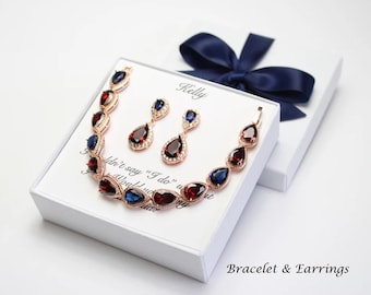 May include: A white gift box with a navy blue ribbon, containing a rose gold bracelet and earrings with red and blue teardrop-shaped stones. The box is labelled "Kelly" and "Bracelet & Earrings".