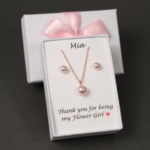 May include: A white gift box with a pink ribbon and a white card inside. The card says "Mia" and "Thank you for being my Flower Girl". The box contains a pink pearl necklace and earrings.