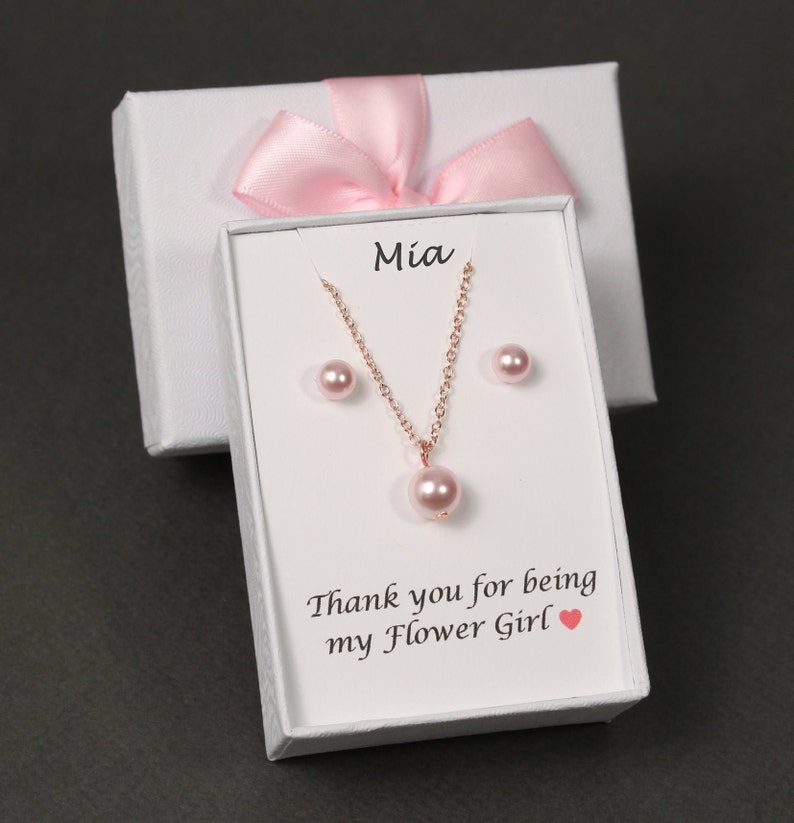 May include: A white gift box with a pink ribbon and a white card inside. The card says "Mia" and "Thank you for being my Flower Girl". The box contains a pink pearl necklace and earrings.