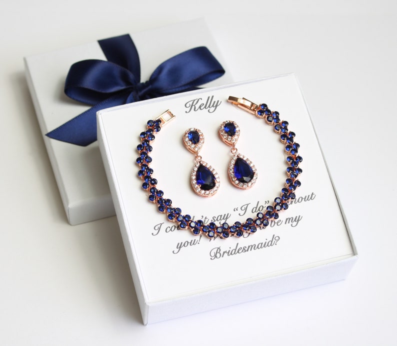 May include: A rose gold bracelet and matching earrings set with dark blue teardrop-shaped stones and clear accents. The jewelry is presented in a white gift box with a navy blue ribbon. Text on the card reads "Kelly, I couldn't say 'I do' without you! Love my Bridesmaid?"