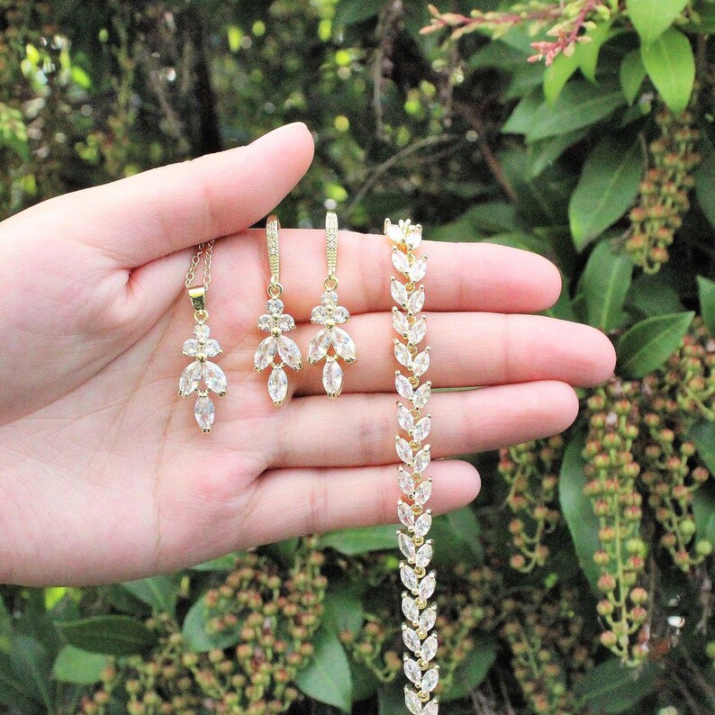 May include: A gold necklace, earrings, and bracelet set with clear marquise-shaped stones. The jewelry is arranged on a hand against a green leafy background.