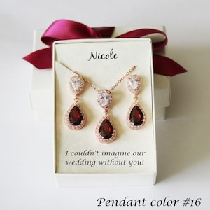 May include: A set of rose gold jewelry with a teardrop pendant and matching earrings. The pendant and earrings feature a large red gemstone surrounded by smaller clear gemstones. The jewelry is in a white box with the name "Nicole" and the message "I couldn't imagine our wedding without you!" The box also has the text "Pendant color #16".