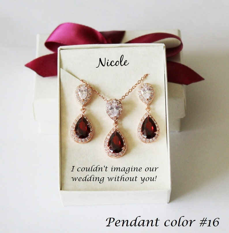 May include: A set of rose gold jewelry with a teardrop pendant and matching earrings. The pendant and earrings feature a large red gemstone surrounded by smaller clear gemstones. The jewelry is in a white box with the name "Nicole" and the message "I couldn't imagine our wedding without you!" The box also has the text "Pendant color #16".