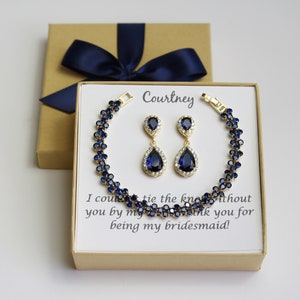 May include: A gold-toned bracelet and matching earrings set with dark blue gemstones and clear accents. The jewelry is presented in a gift box with a navy blue bow. The box contains a card with the name "Courtney" and a message.