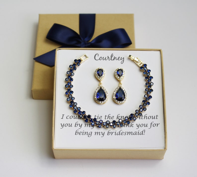 May include: A gold-toned bracelet and matching earrings set with dark blue gemstones and clear accents. The jewelry is presented in a gift box with a navy blue bow. The box contains a card with the name "Courtney" and a message.