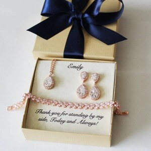 Puede incluir: Una caja de regalo con una cinta azul marino y un mensaje personalizado para Emily. Dentro de la caja hay un juego de joyas de oro rosa con un collar, pendientes y una pulsera. Las joyas presentan piedras en forma de l&aacute;grima con un halo de piedras m&aacute;s peque&ntilde;as.