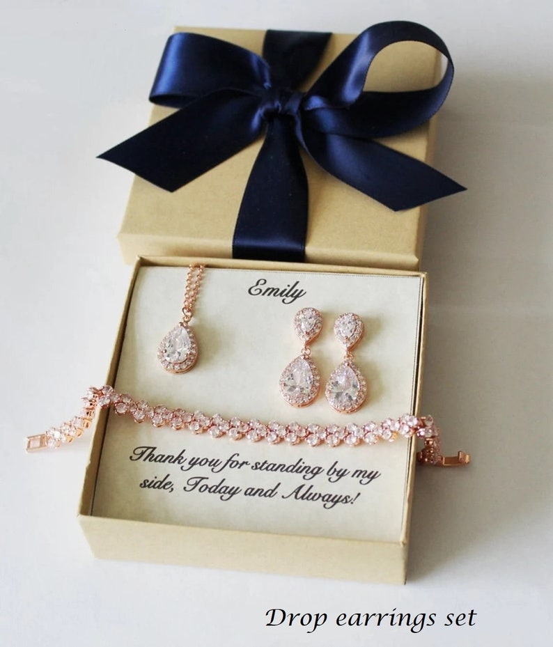 Puede incluir: Una caja de regalo con una cinta azul marino y un mensaje personalizado para Emily. Dentro de la caja hay un juego de joyas de oro rosa con un collar, pendientes y una pulsera. Las joyas presentan piedras en forma de l&aacute;grima con un halo de piedras m&aacute;s peque&ntilde;as.