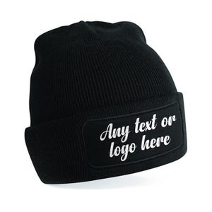 May include: Black knitted beanie hat with a rectangular patch. The patch displays the text "Any text or logo here" in white script. The hat is made of a ribbed knit material and has a folded brim.