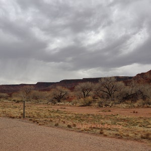 K&ouml;nnte beinhalten: Landschaft mit einer Stra&szlig;e, die zu einer Bergkette unter einem bew&ouml;lkten Himmel f&uuml;hrt. Der Vordergrund zeigt trockene Vegetation und eine unbefestigte Stra&szlig;e. Der Text "Live With Beauty" ist unten rechts sichtbar.