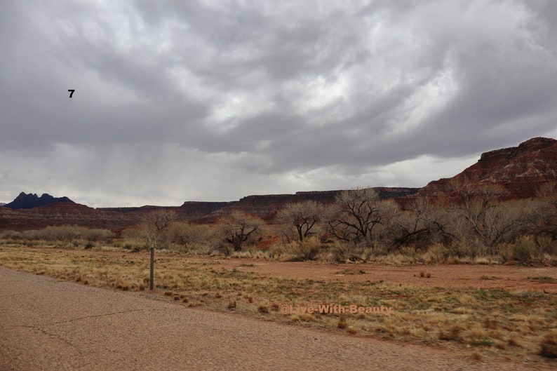 K&ouml;nnte beinhalten: Landschaft mit einer Stra&szlig;e, die zu einer Bergkette unter einem bew&ouml;lkten Himmel f&uuml;hrt. Der Vordergrund zeigt trockene Vegetation und eine unbefestigte Stra&szlig;e. Der Text "Live With Beauty" ist unten rechts sichtbar.