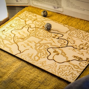 May include: A wooden map with a detailed, burnt-in design of a fantasy landscape, featuring rivers, mountains, and forests. Three metallic dice are placed on the map and surrounding surface. The map is resting on a textured, golden-yellow fabric.