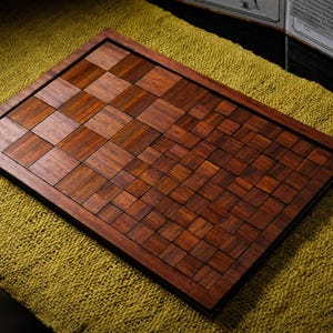 May include: A handcrafted wooden chessboard with a dark brown frame and a checkerboard pattern of alternating light and dark brown squares. The board is set on a textured, golden-yellow surface.
