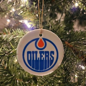 Op de afbeelding: Een ronde, witte keramische kerstbal met het logo van de Edmonton Oilers. Het logo bevat een blauwe cirkel met het woord "OILERS" in blauwe letters en een oranje oliedruppel. De bal hangt aan een gouden draad.
