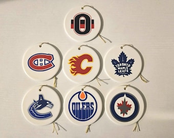 Könnte beinhalten: Eine Sammlung weißer Keramik-Ornamente mit Logos verschiedener Eishockeyteams, darunter die Montreal Canadiens, Calgary Flames und Toronto Maple Leafs. Jedes Ornament hat eine goldene Schnur zum Aufhängen.