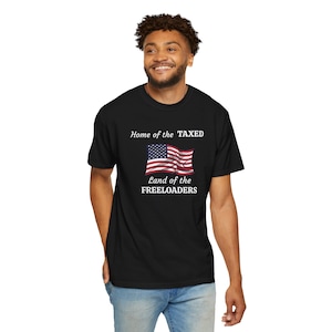 May include: Black t-shirt featuring the American flag with the text "Home of the TAXED, Land of the FREELOADERS." The design includes a waving flag in red, white, and blue. The shirt is a classic crew neck style.
