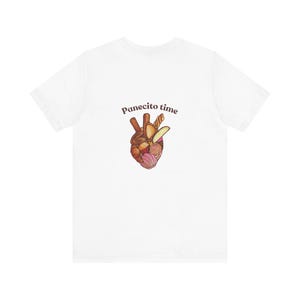 Puede incluir: Camiseta blanca con un diseño en forma de corazón compuesto por varios productos de panadería en tonos marrones, rosas y amarillos. El texto "Panecito time" está encima del corazón. La camiseta es de un material suave y cómodo.