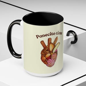 Puede incluir: Taza de cerámica blanca con interior y asa negros. La taza presenta el texto "Panecito time" sobre una ilustración en forma de corazón de varios pasteles en tonos marrón, rosa y amarillo.