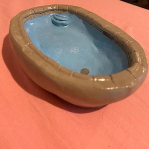 May include: A miniature, hand-painted ceramic pool with a light blue interior and a tan exterior. The pool features a small set of steps and is oval-shaped. The rim of the pool has a textured, stone-like appearance.
