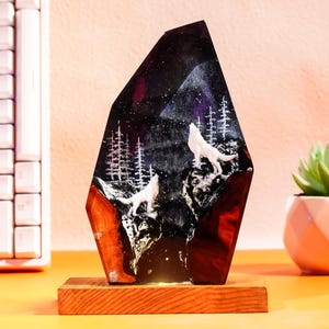 Könnte beinhalten: Dekorative Harz-Nachtlampe mit einer Berglandschaft mit zwei weißen Wölfen, die den Mond anheulen. Das Nachtlicht steht auf einem Holzsockel und hat einen dunklen, sternenübersäten Hintergrund mit weißen Bäumen.