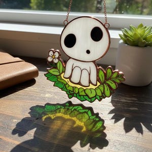 May include: Stained glass suncatcher featuring a white spirit character with black eyes and a surprised expression, nestled in green leaves. The suncatcher hangs from a copper chain, casting a colorful shadow. A small white flower is visible.