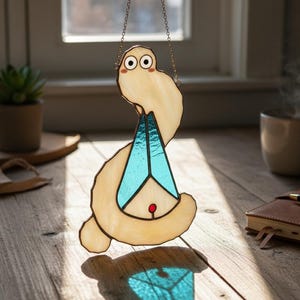 Puede incluir: Un atrapasoles de vidriera con forma de criatura caprichosa con cuerpo beige claro, detalles geométricos azules y un punto rojo. Cuelga de una cadena y proyecta una sombra azul. Cerca de una ventana.