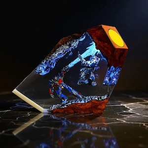 May include: A resin sculpture of two robots in action, with blue and red detailing. The sculpture is encased in clear resin, with a wooden and yellow accent on the top. The base is dark, and the sculpture is angled. The measurements are in centimeters.