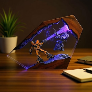 Puede incluir: Escultura decorativa de resina que representa una escena de batalla entre dos figuras de robots, iluminada por una luz púrpura. La escultura está encerrada en un marco de madera con una parte frontal transparente, creando un efecto 3D. Las figuras son naranjas y azules.