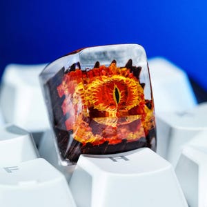 May include: A custom keycap with a fiery eye design set in clear resin. The keycap, with its orange and yellow eye detail, sits on a white keyboard. A unique accessory for mechanical keyboards, it measures approximately 2 cm in length.