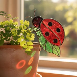 Pode incluir: Uma decoração de joaninha em vitral com corpo vermelho e pintas pretas, pousada numa folha verde. A joaninha está colocada perto de uma planta em vaso com folhas verdes. O vaso é castanho e está perto de uma janela.