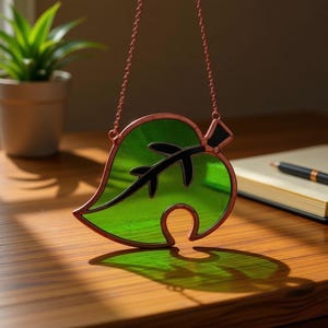 May include: A stained glass leaf with a copper-colored frame and chain. The leaf is a vibrant green with black accents, casting a shadow on a wooden surface. The design is reminiscent of a video game.