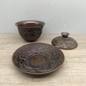May include: A bronze-colored tea set with intricate dragon relief carvings. The set includes a small bowl, a saucer, and a lid. The pieces are arranged on a light-colored wooden surface.